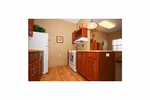 Interior view of a kitchen in a senior living facility featuring wooden cabinets, a white refrigerator, a white stove with an overhead range hood, and a wooden floor. The kitchen opens into a living area with a window and a beige wall.