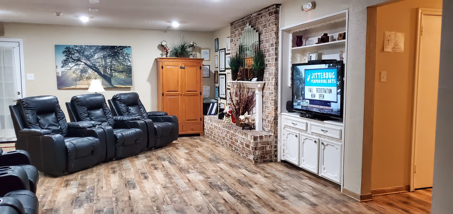 A cozy living room with four black leather recliners arranged in a row facing a brick fireplace and a white built-in entertainment center with a TV displaying a sign for Jitterbug Performing Arts fall registration. The room has wood flooring, a wooden cabinet, framed pictures on the walls, and a large tree artwork above a lamp on a side table.