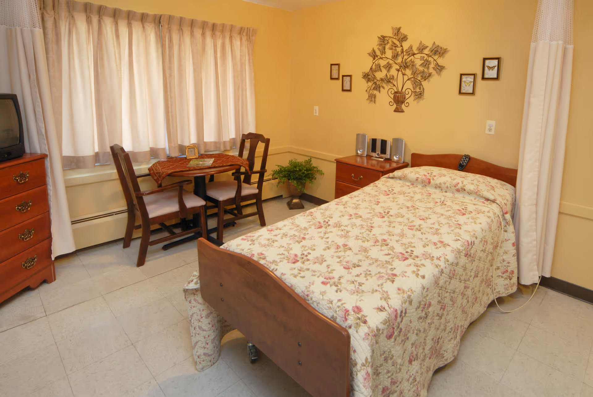 A cozy bedroom with a single bed covered in a floral bedspread. Next to the bed is a wooden nightstand with a small stereo system. There is a small table with two chairs near a window with beige curtains. The walls are painted yellow and decorated with framed pictures and a metal wall art piece shaped like a tree with butterflies. A wooden dresser with a small TV on top is visible on the left side of the room.