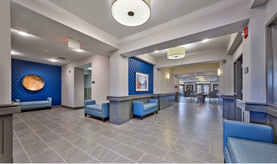 Modern senior living lobby with blue seating, decorative wall panels and artwork, and a long tiled hallway leading to a dining area.