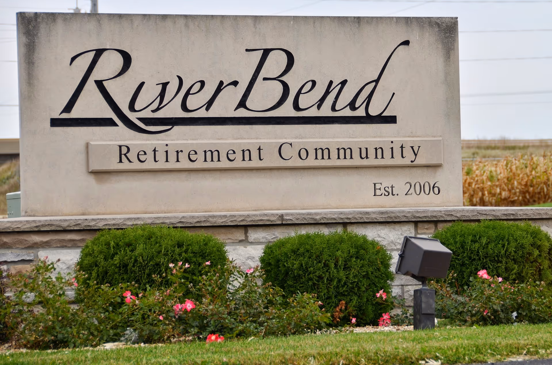 Stone entrance sign reading "RiverBend Retirement Community Est. 2006" with shrubs and flowers in front.