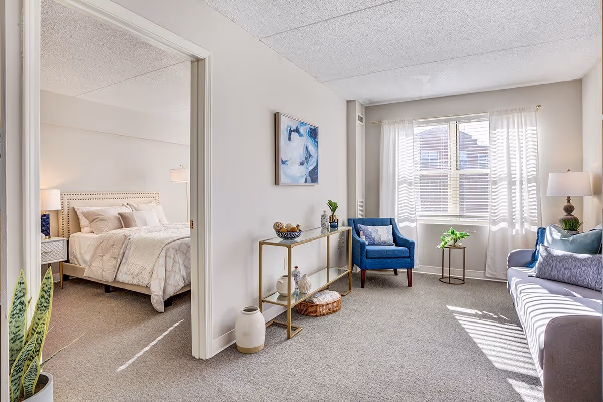Bright living room with a blue armchair and sofa, a console table, and an open doorway showing a furnished bedroom.