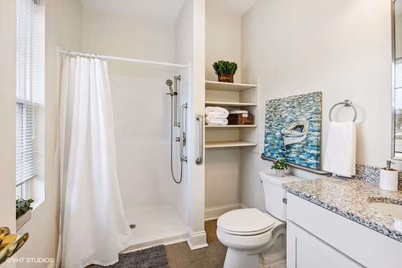 A bright bathroom featuring a walk-in shower with a white curtain, a toilet, and a granite countertop with a sink. There are built-in shelves holding folded towels and a potted plant. A painting of a boat on water hangs above the toilet, and a towel ring with a white towel is mounted on the wall next to the sink.