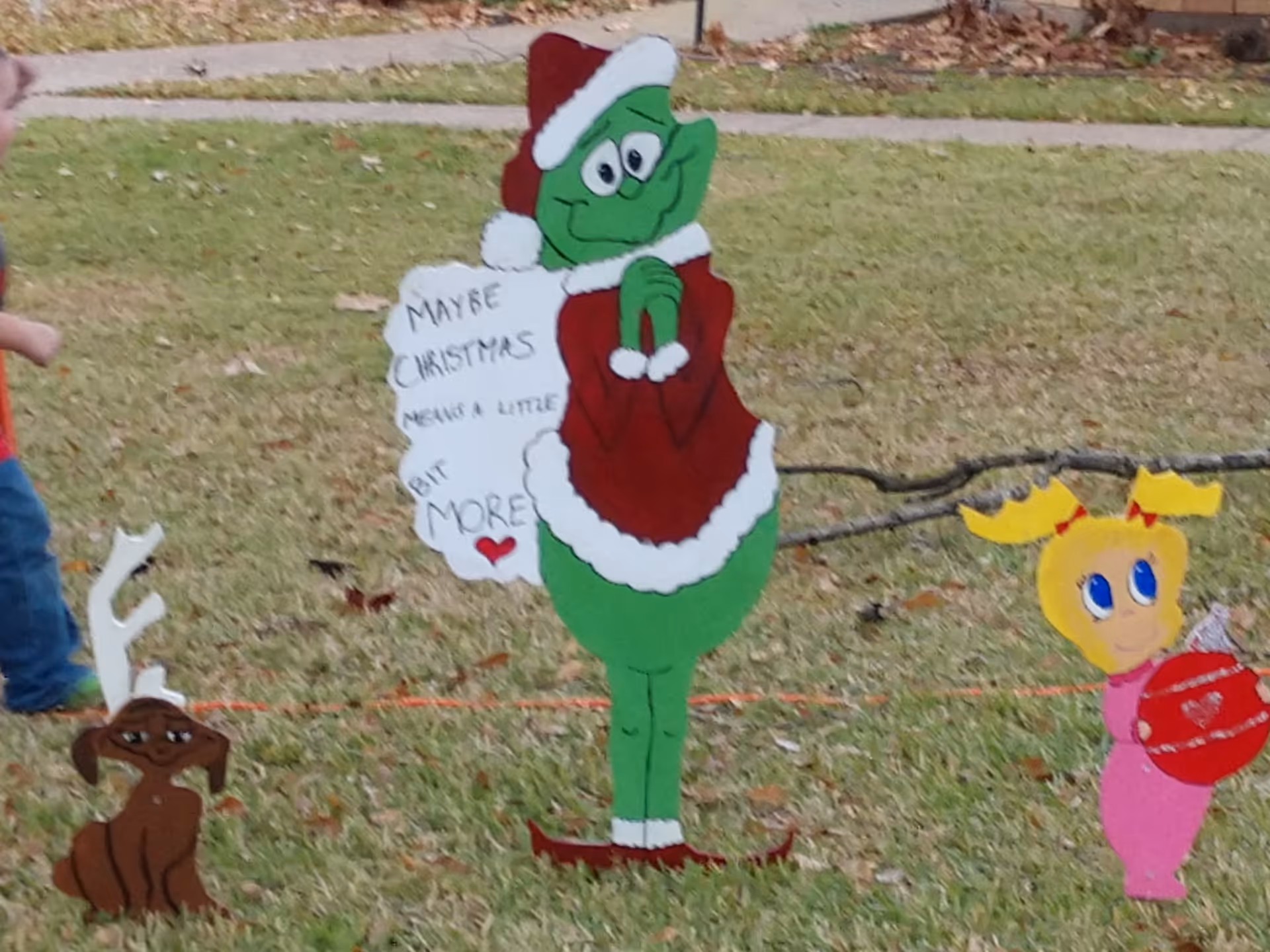 Outdoor Christmas decorations on grass including a green character dressed in a red Santa outfit holding a sign that says 'Maybe Christmas means a little bit more', a small brown dog with white antlers, and a yellow character with antlers holding a red ornament.