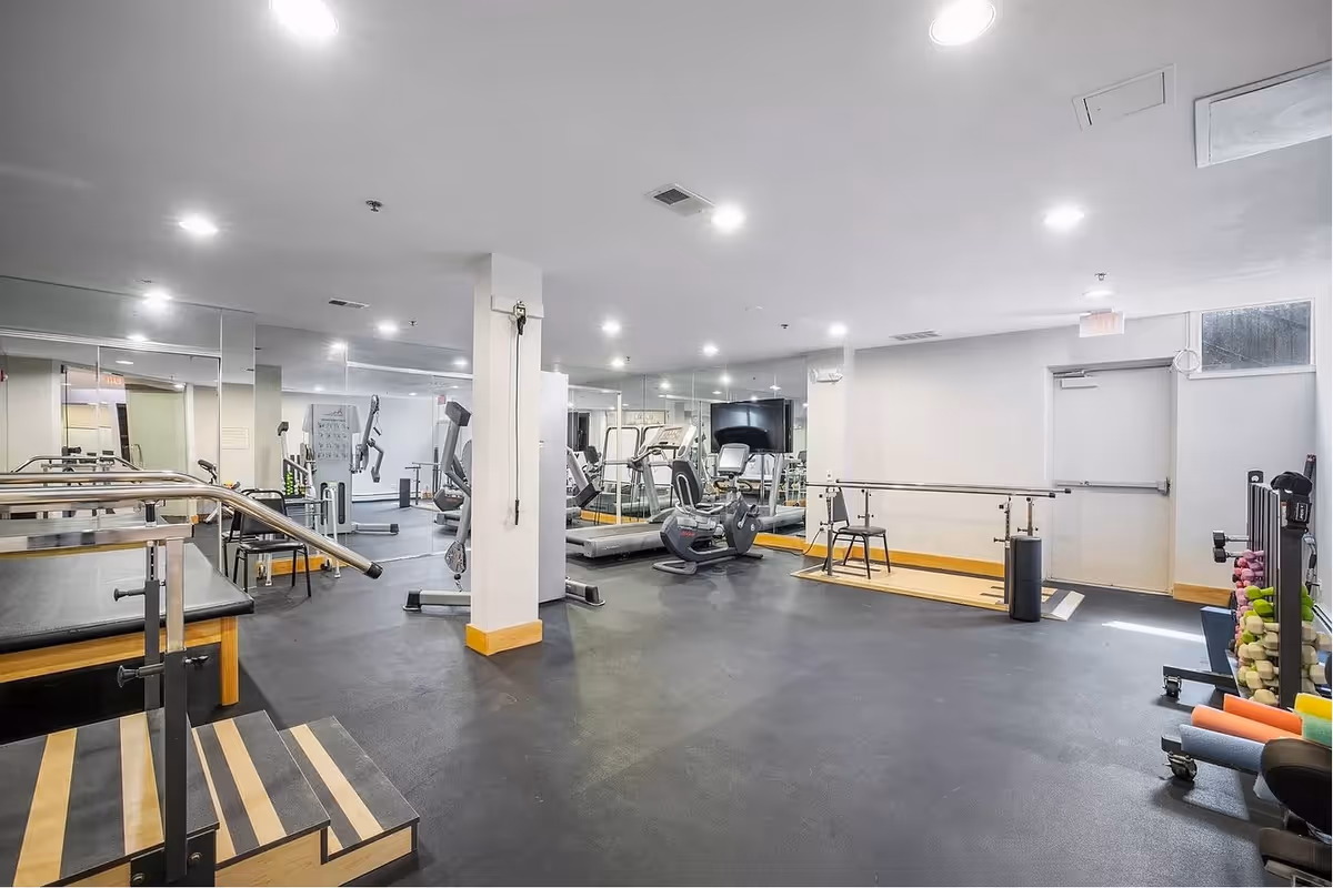 A spacious fitness room with exercise equipment including treadmills, stationary bikes, hand weights, and parallel bars. The room has mirrors on the walls, bright overhead lighting, and a black rubber floor with wooden accents. There is a chair and a small platform with steps for exercise or physical therapy.