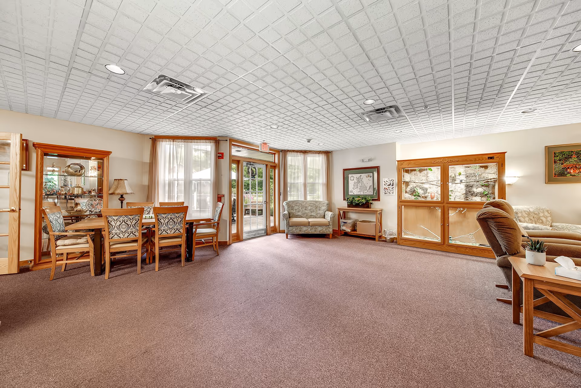 A spacious senior living common area with carpeted floor, a dining table with six chairs on the left, a loveseat near large windows with sheer curtains, and a comfortable armchair with a side table on the right. The room has a white patterned ceiling with recessed lighting and wooden framed windows and doors leading outside.