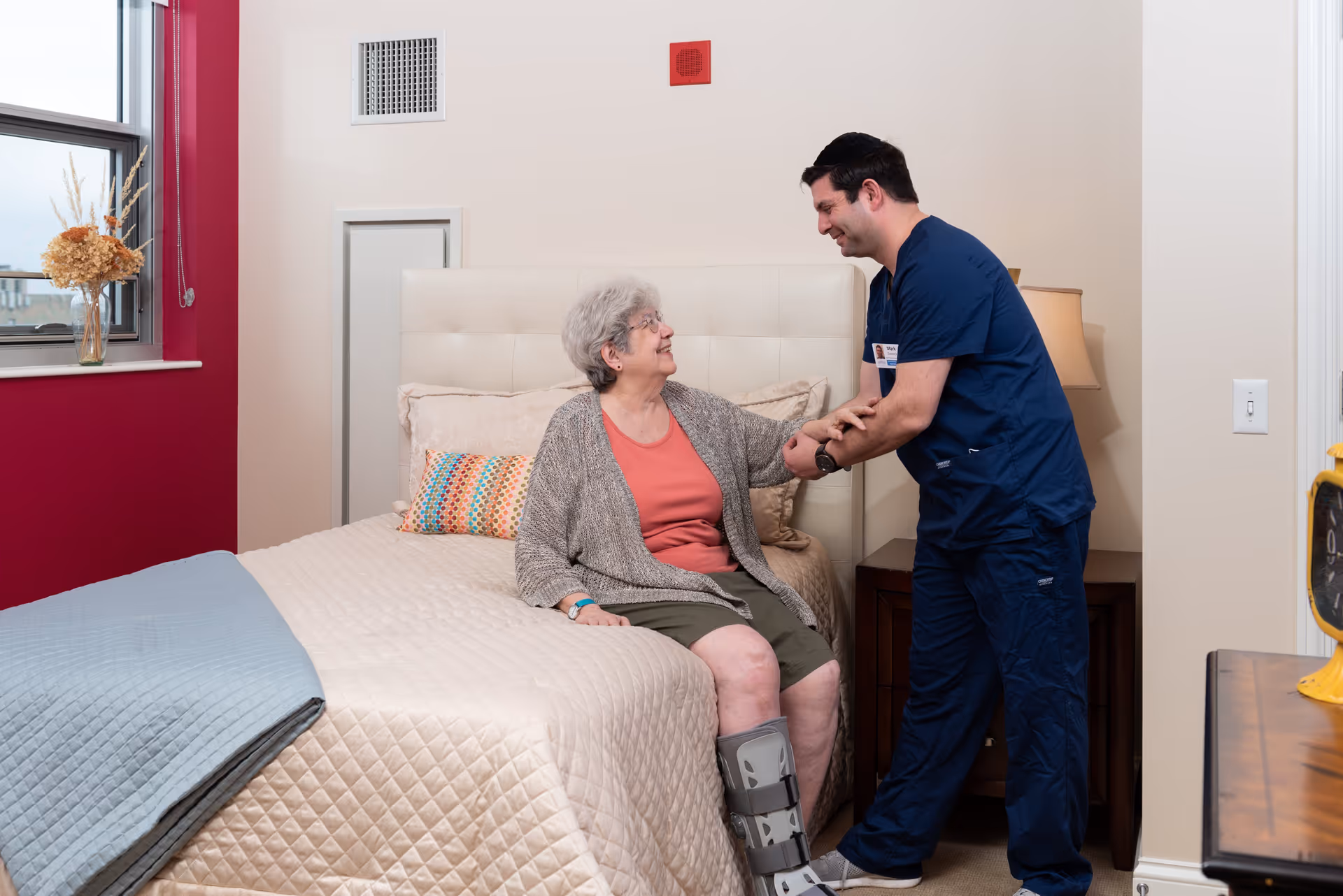 An elderly woman with a leg brace sitting on a bed in a bedroom, smiling and holding hands with a male healthcare worker in navy scrubs who is standing beside her. The room has a beige and red color scheme with a window, a vase with dried flowers, and a bedside table with a lamp.