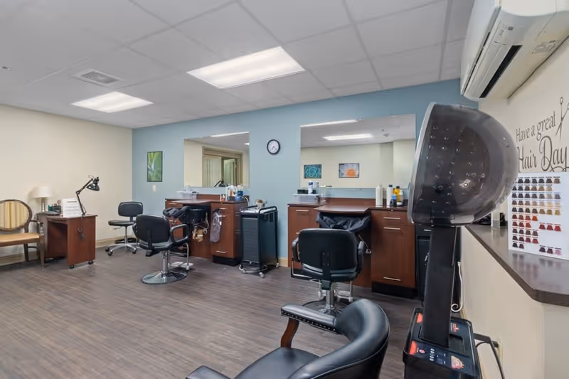 Interior view of a hair salon area in a senior living facility with salon chairs, mirrors, hair care products, a hair dryer station, and a sign on the wall that says 'Have a great Hair Day'.