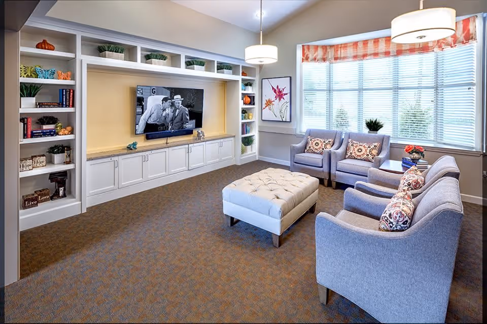 A cozy living room with a large window covered by blinds and a valance. The room features a built-in white entertainment center with shelves holding decorative items and a flat-screen TV showing a black and white movie. There are four upholstered armchairs with patterned cushions arranged around a tufted ottoman in the center. Two pendant lights hang from the ceiling.