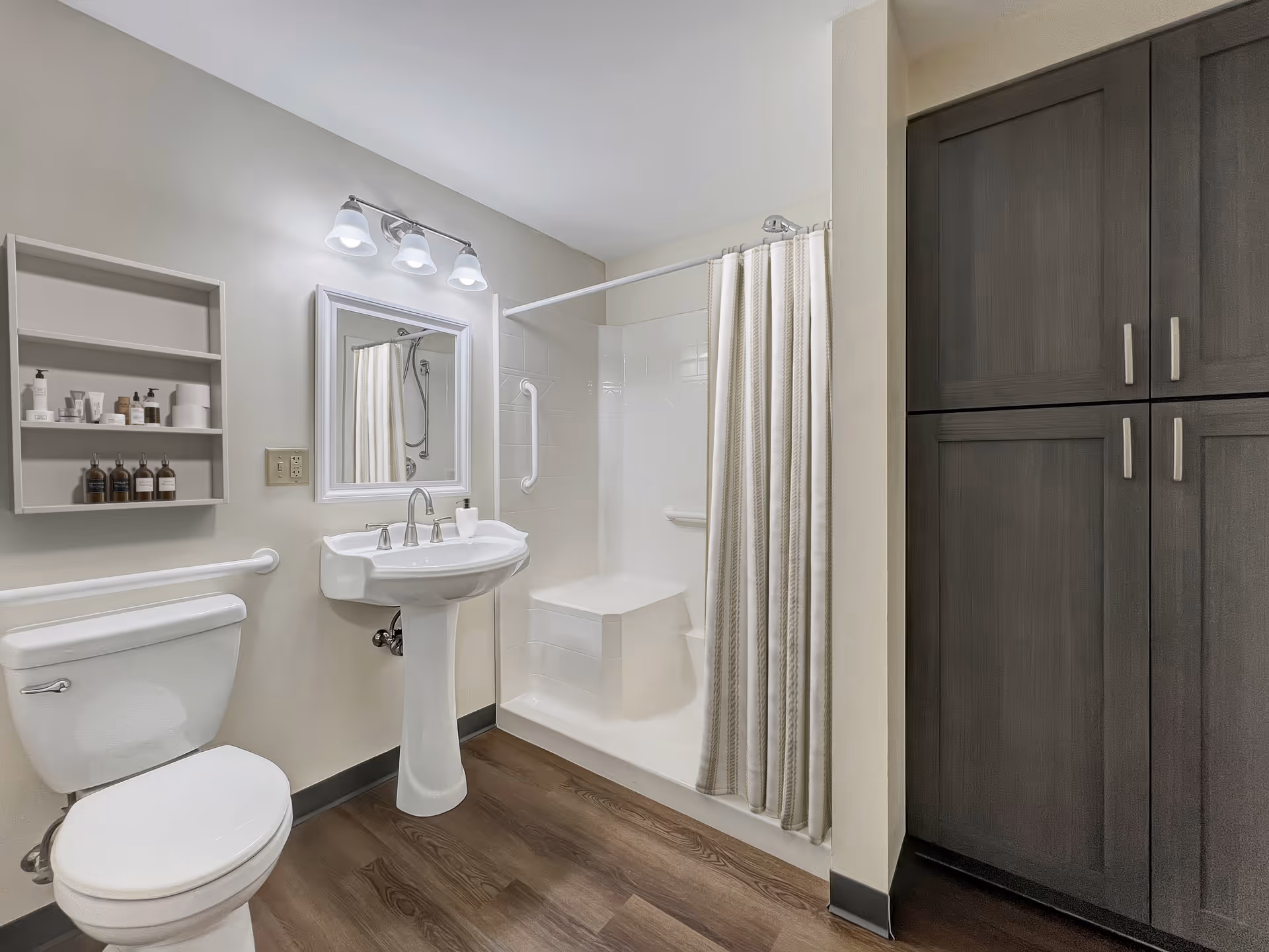 A clean and modern bathroom featuring a white toilet, a pedestal sink with a mirror and three-light fixture above it, a walk-in shower with a built-in seat and grab bars, a beige shower curtain, wall-mounted shelves with toiletries, and dark wood cabinets on the right side. The floor has wood-like vinyl flooring.