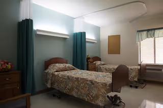 A hospital-style room with two beds separated by green privacy curtains. Each bed has a floral patterned bedspread. There is a wooden nightstand with flowers between the beds, a window with blinds and a valance, and a wooden dresser on the left side of the room.