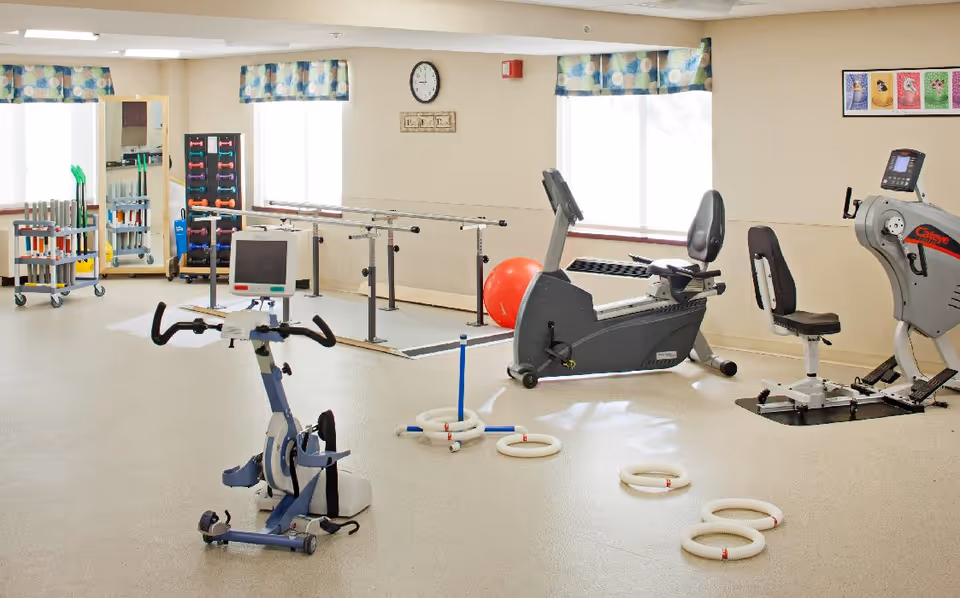 A bright exercise room with various fitness equipment including a stationary bike, recumbent bike, elliptical machine, parallel bars for walking exercises, a red exercise ball, and a ring toss game on the floor. The room has large windows with patterned valances, a clock on the wall, and racks holding dumbbells and other exercise tools.