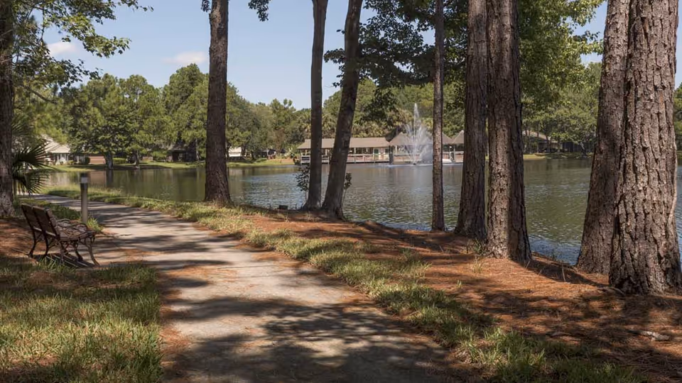 A peaceful outdoor scene at Cypress Village featuring a paved walking path alongside a lake with tall trees providing shade. There is a bench on the left side of the path, and a water fountain is visible in the lake with buildings in the background under a clear blue sky.