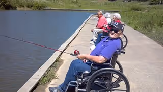 Three elderly individuals sitting in wheelchairs along a concrete path by a body of water, fishing with fishing rods on a sunny day.