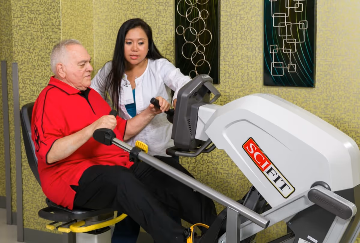 An elderly man in a red shirt is using a seated exercise machine while a female healthcare worker in a white coat assists and supports him. The background features a green patterned wall with decorative metal wall art.