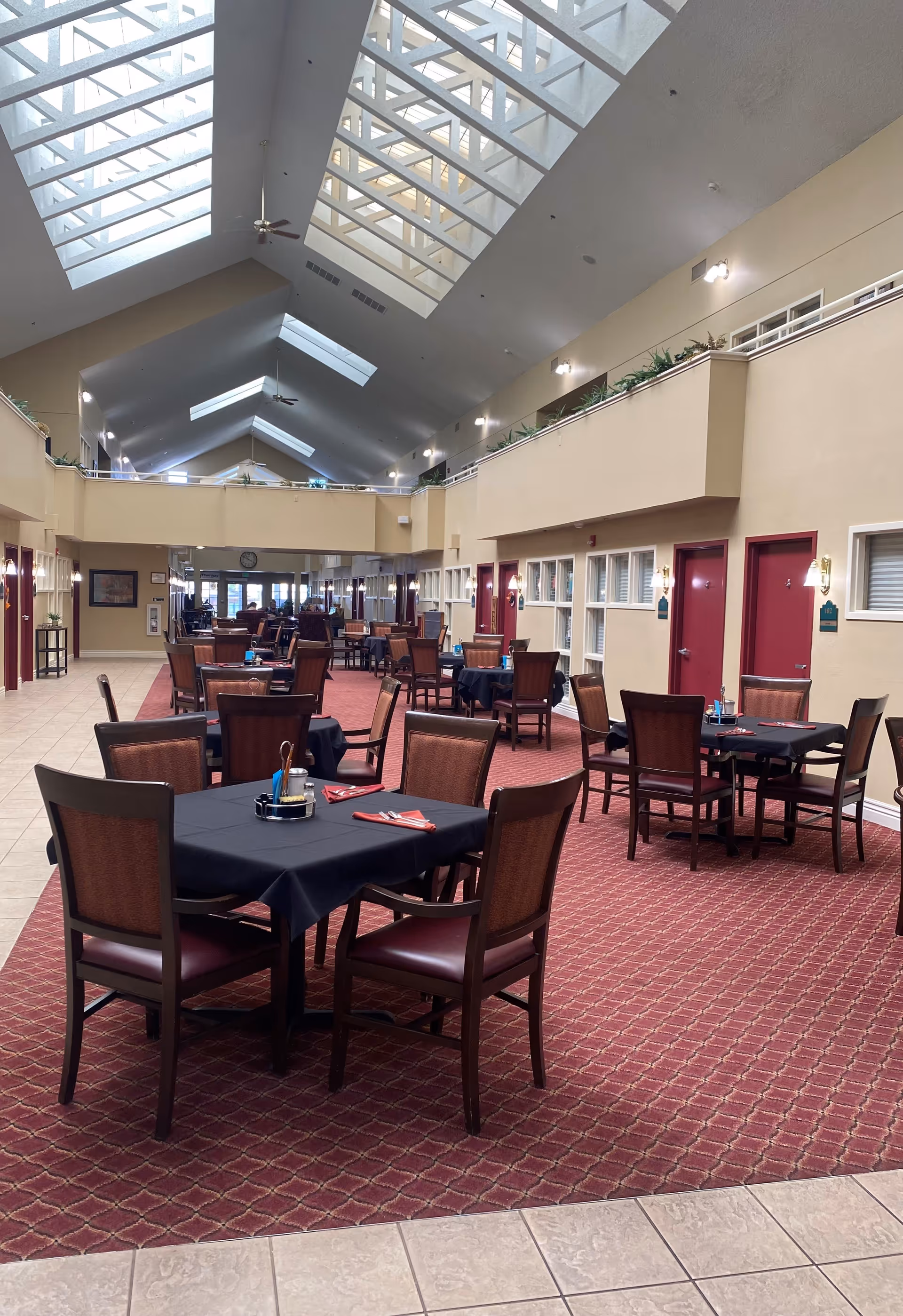 Interior view of a spacious dining area with multiple tables covered with black tablecloths and surrounded by wooden chairs with cushioned seats. The room features a high ceiling with large skylights allowing natural light to fill the space. The floor is carpeted in a red patterned design, and there are beige walls with red doors and windows along one side. The area appears clean and well-lit, designed for communal dining.