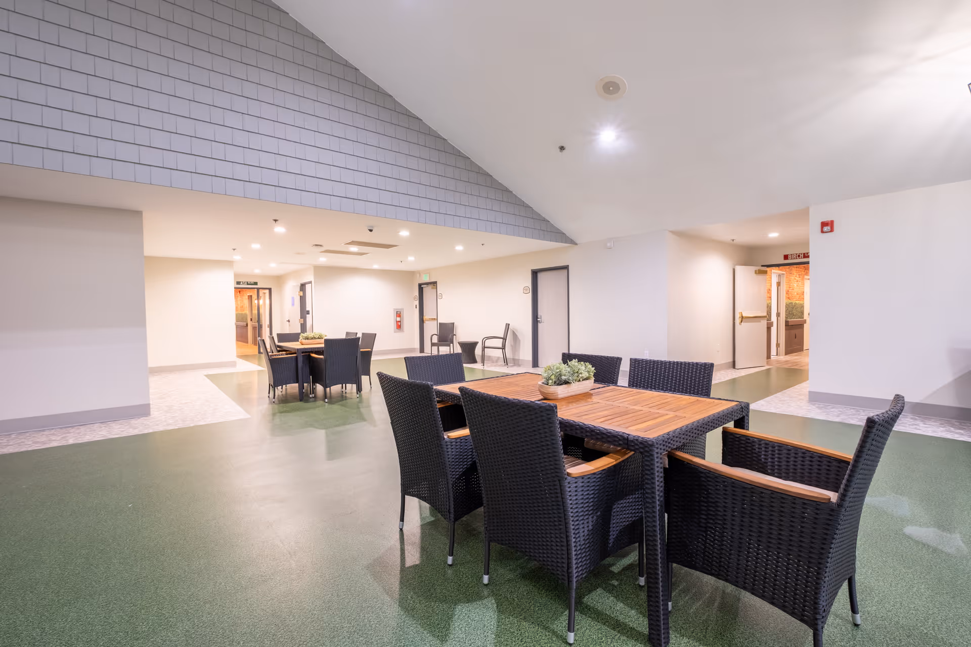 Indoor common area with green flooring and white walls featuring two sets of dark wicker tables and chairs with wooden tabletops. The ceiling is white with recessed lighting, and there are doors and hallways visible in the background.