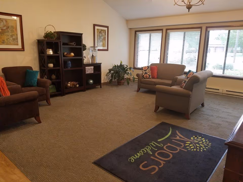 A cozy living room area with two gray upholstered armchairs and a matching loveseat, each adorned with colorful throw pillows. There is a brown armchair with a teal pillow and an orange blanket on the left side. A dark wooden shelving unit and cabinet stand against the wall, decorated with various items and a plant. Large windows with blinds let in natural light. A dark welcome mat with the words 'Arbors Welcome' and a tree logo is placed on the carpeted floor near the entrance.