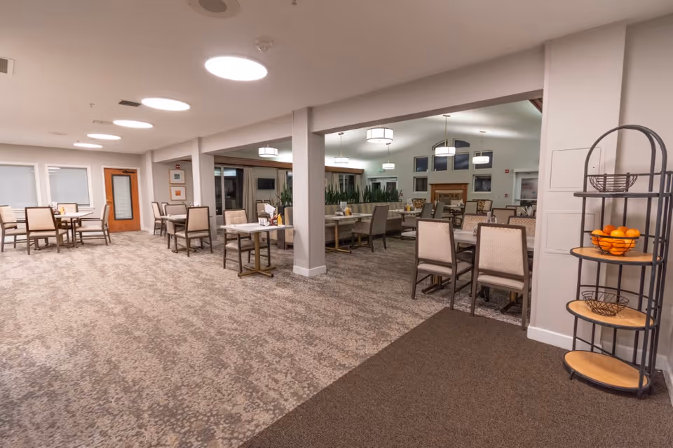 A spacious dining room in a senior living facility with multiple tables and chairs arranged neatly. The room features carpeted floors, modern ceiling lights, large windows, and a fireplace in the background. A metal rack with baskets and a bowl of oranges is visible on the right side.