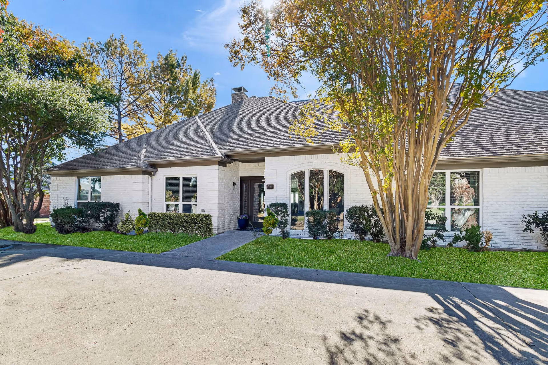 Exterior view of a single-story white brick building with a dark shingled roof, surrounded by green grass, bushes, and trees under a clear blue sky.