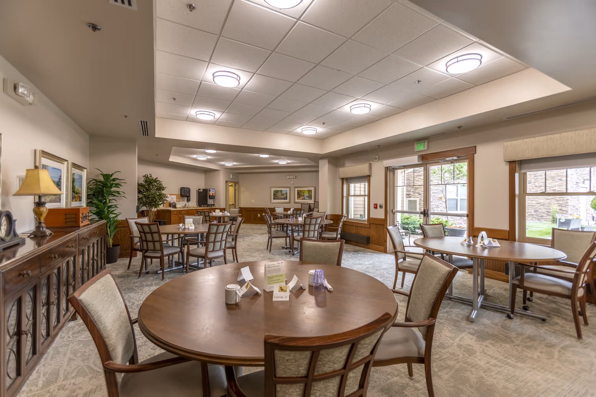A spacious dining room in a senior living facility with multiple round tables and chairs arranged neatly. The room has large windows allowing natural light, beige walls, carpeted floors, ceiling lights, and decorative plants. A sideboard with a lamp and framed pictures is visible on the left side.