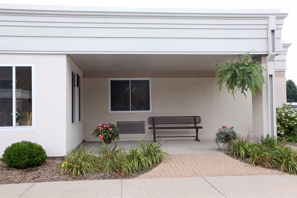Covered front entrance with a metal bench, potted flowers, hanging fern, and landscaped plant beds.