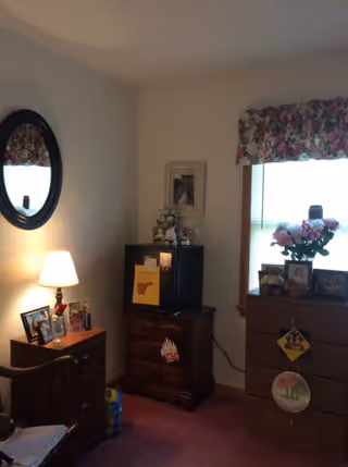 A cozy bedroom corner with wooden furniture including a dresser with framed photos and a lamp, a small cabinet with a mini fridge on top, a window with floral valance, and decorative items such as flowers and wall hangings.