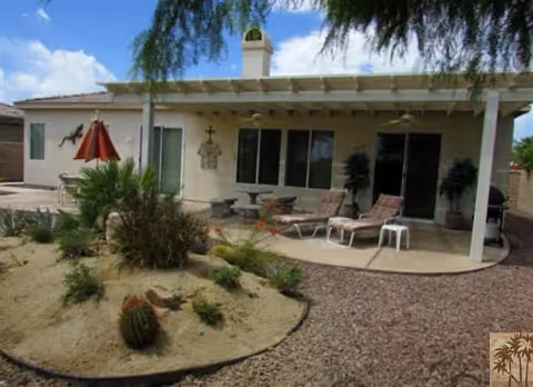 Backyard patio area of a residential care facility with two lounge chairs, a small white side table, potted plants, and a covered patio attached to a beige building. The yard features desert landscaping with gravel and various cacti and shrubs under a partly cloudy sky.