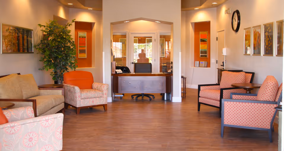 A well-lit reception area with a wooden desk and office chair in the center background. The room features comfortable seating including beige and patterned armchairs with orange cushions, a beige sofa, wooden side tables, a large potted plant, colorful framed artwork on the walls, and a clock. The floor is wooden and the walls are painted in neutral tones.