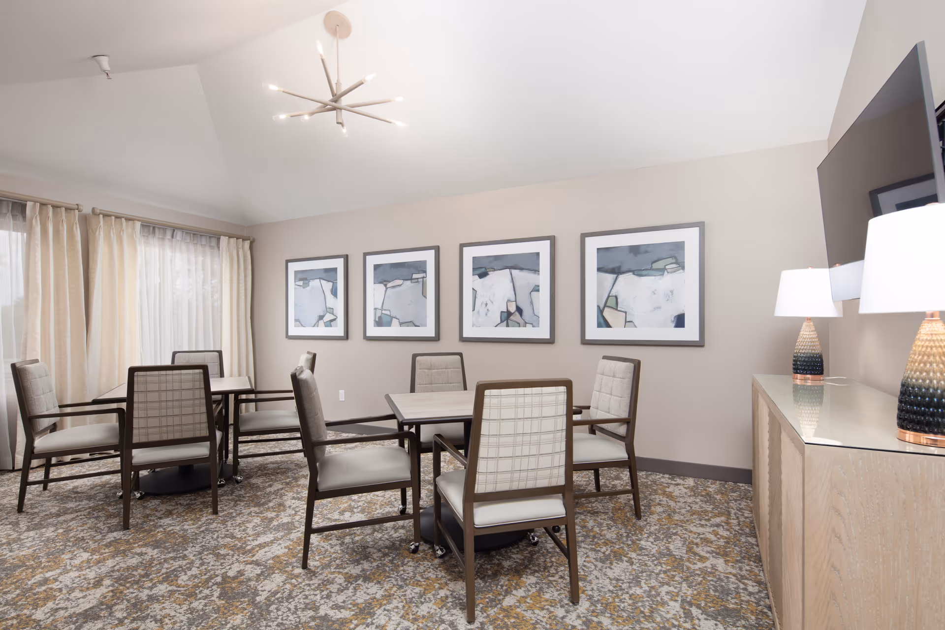 A well-lit room with two square tables, each surrounded by four cushioned chairs with wooden frames. The room features a patterned carpet, beige walls adorned with four abstract art paintings, a modern chandelier hanging from the ceiling, and a sideboard with two decorative lamps and a mounted flat-screen TV.