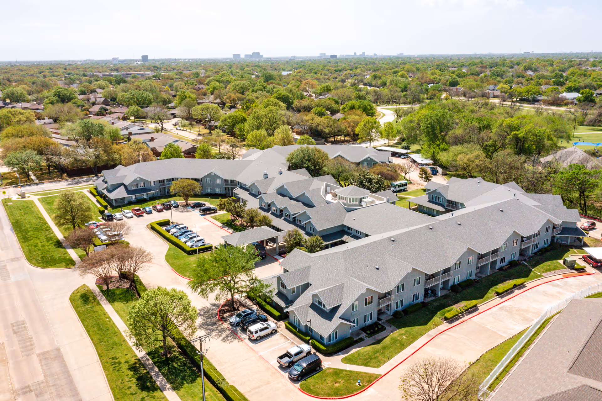 Aerial view of Solstice Senior Living at Plano, showing a large senior living facility with multiple connected buildings surrounded by trees and greenery. There are parking lots with several cars parked, and the facility is located in a suburban area with residential houses and roads nearby.