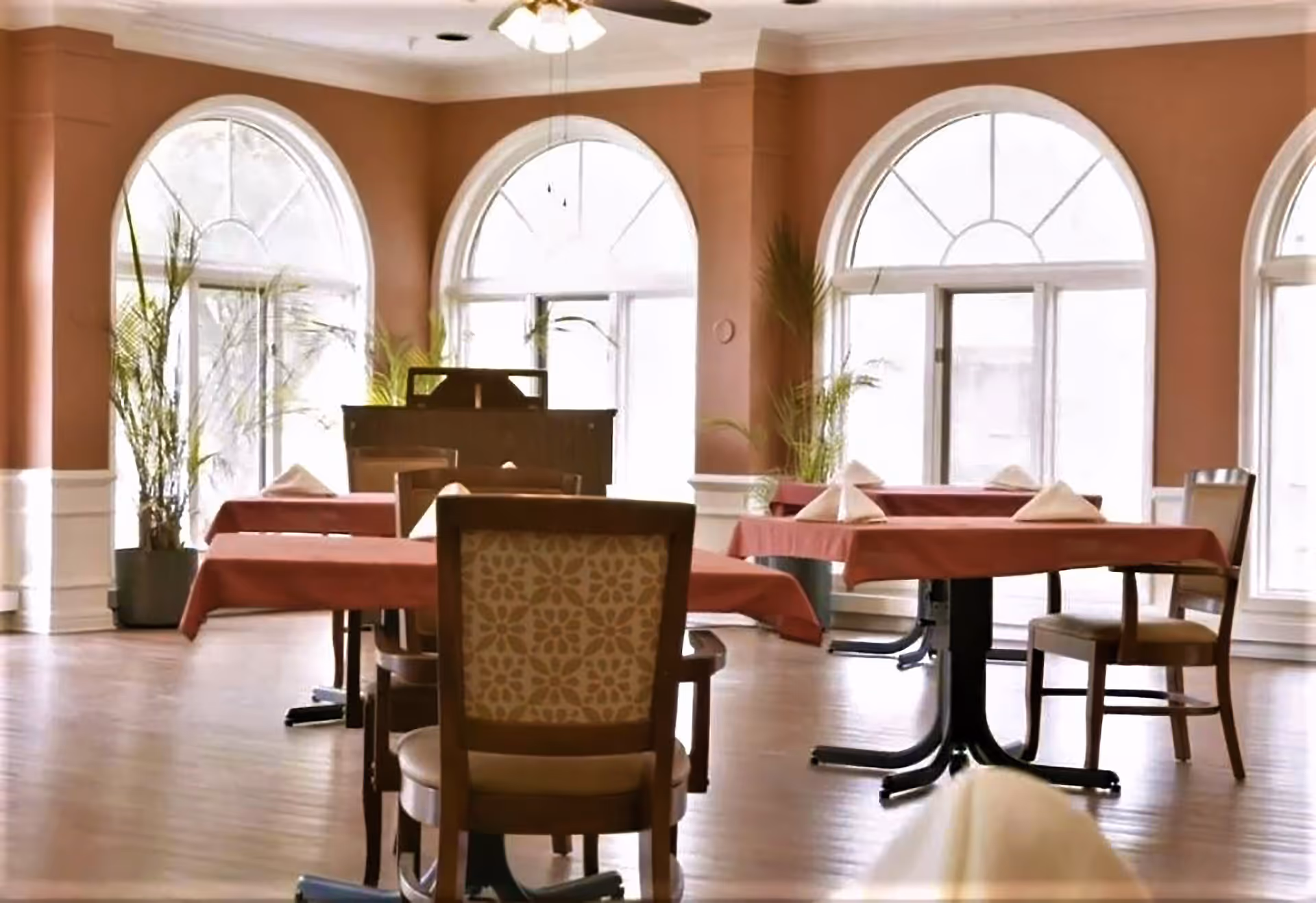 Sunlit dining room with tables set with red tablecloths and chairs arranged near large arched windows and potted plants.