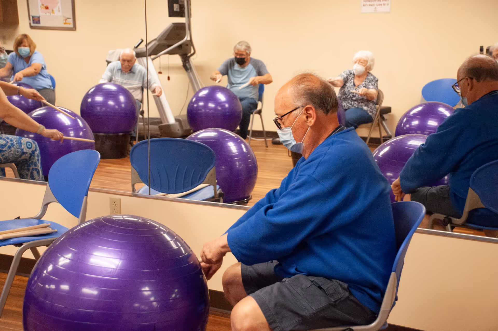 A group of elderly individuals wearing masks participate in a seated exercise class using large purple exercise balls in a room with wooden floors and a mirrored wall.