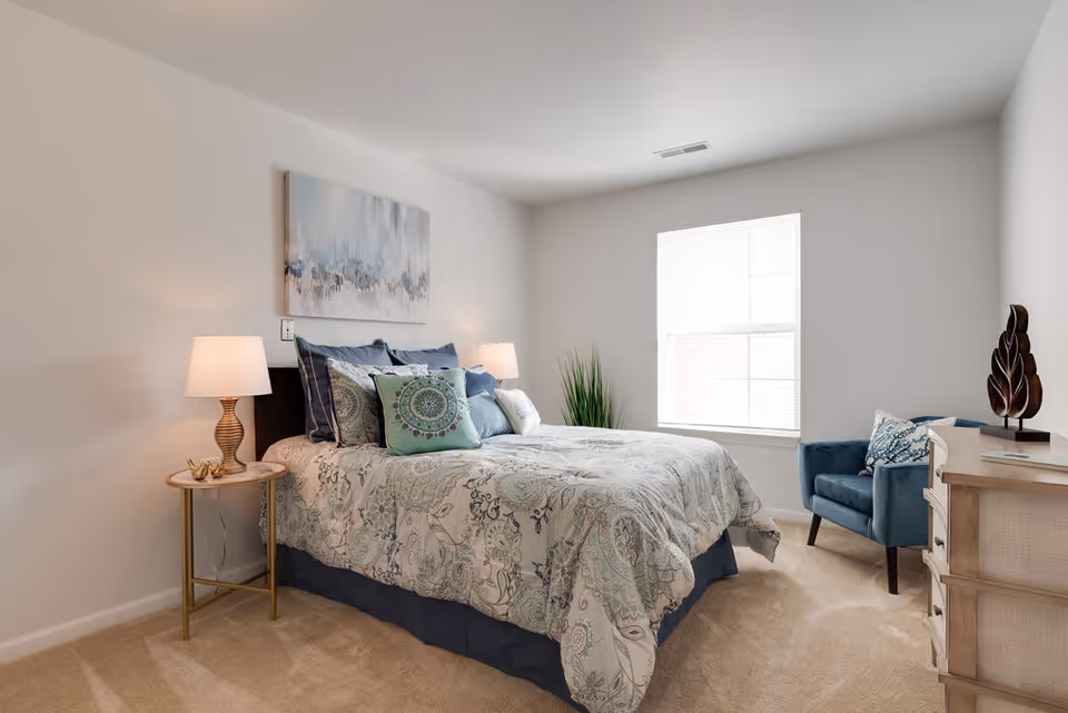 A bright bedroom with a large bed covered in a patterned comforter and multiple pillows. There are two bedside tables with lamps on either side of the bed. A blue armchair is placed near a window with white blinds, and a wooden dresser with decorative items is on the right side of the room. The walls are painted white, and a piece of abstract artwork hangs above the bed.