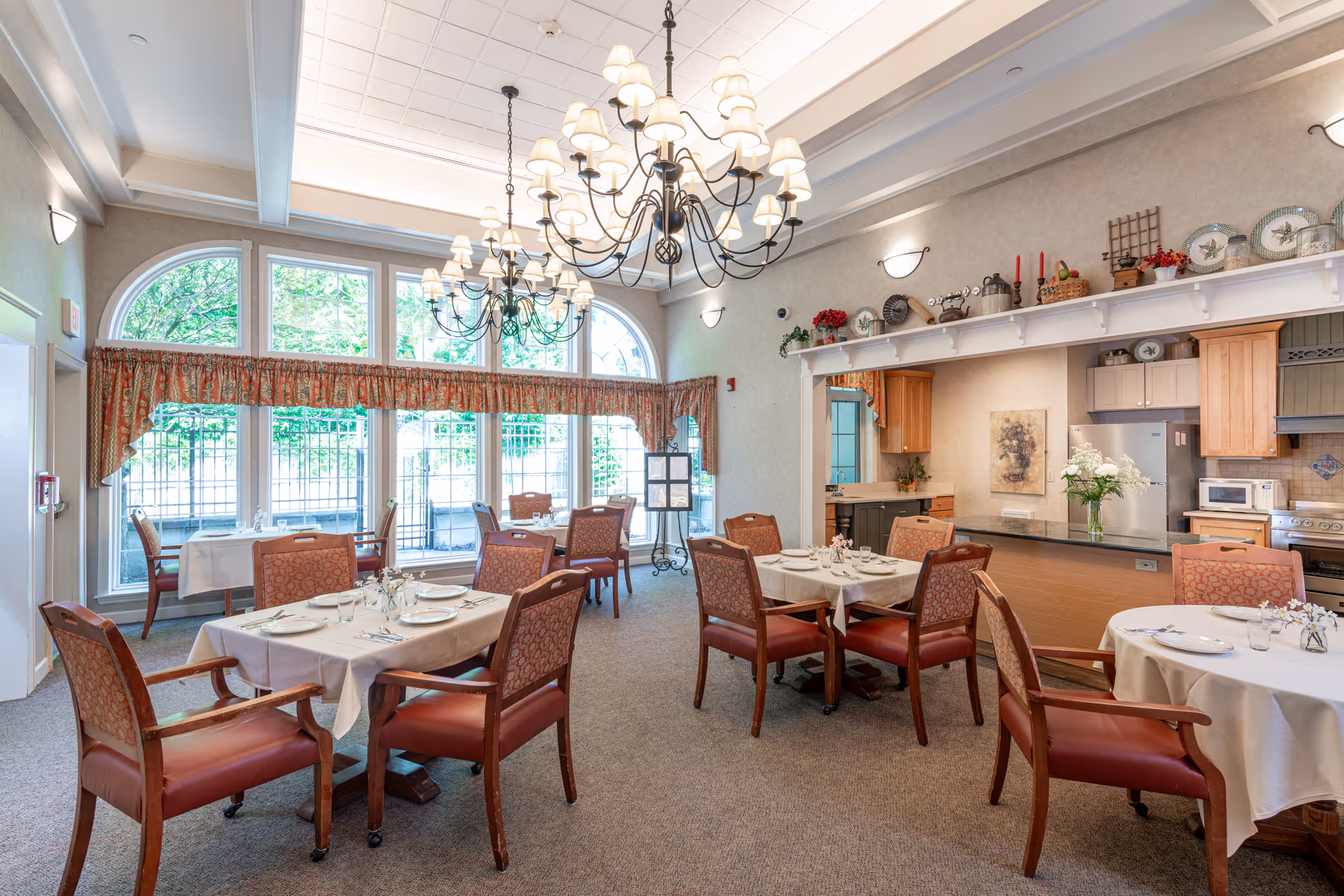 Bright dining room with multiple set tables and chairs, large chandeliers, and a serving counter with a kitchen area.