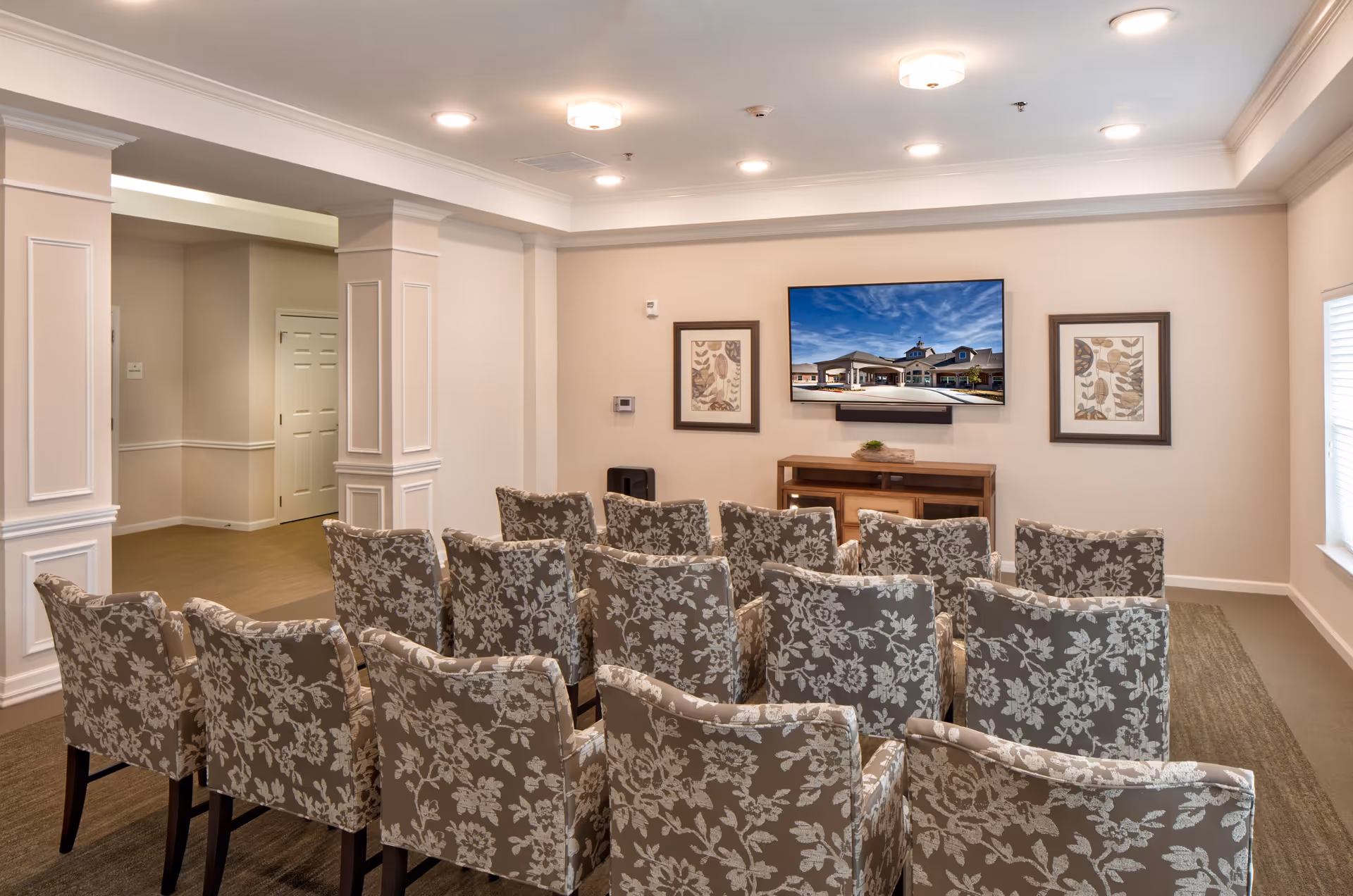 A small theater or media room with three rows of floral-patterned upholstered chairs facing a wall-mounted flat-screen TV. The TV displays an image of a building exterior. The room has beige walls, white trim, recessed ceiling lights, and two framed botanical prints on either side of the TV. There is a wooden console table beneath the TV and a window with blinds on the right side.