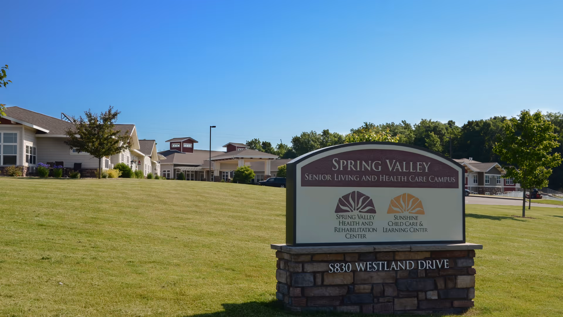 Exterior view of Spring Valley Senior Living and Health Care Campus showing a large green lawn, a stone sign with the facility name and address, and buildings in the background under a clear blue sky.