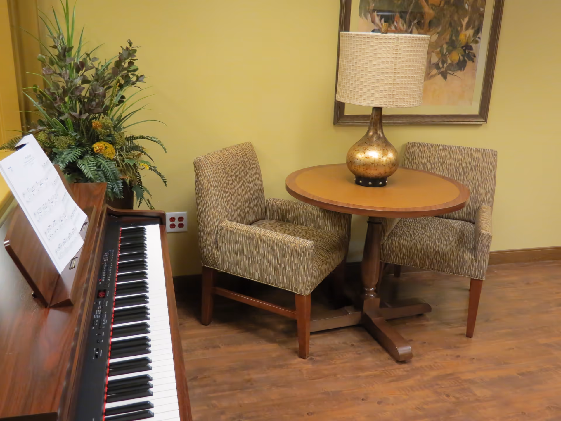 A cozy corner in a memory care facility featuring a wooden piano with sheet music, a round wooden table with a decorative lamp, two upholstered chairs, a framed floral painting on the wall, and a large plant in the corner.