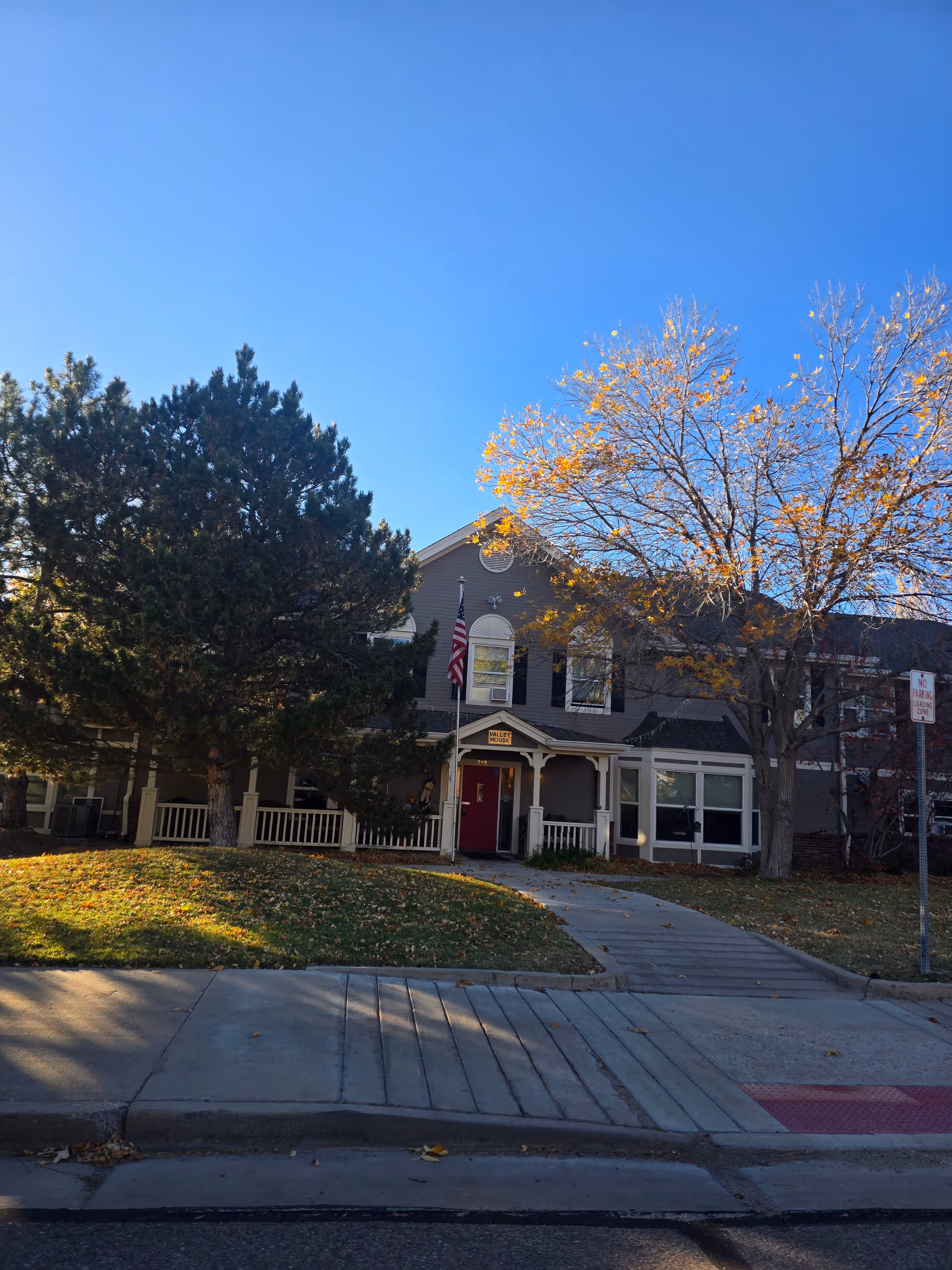 Front exterior of a two-story assisted living building with a porch, American flag, trees, and a sidewalk.