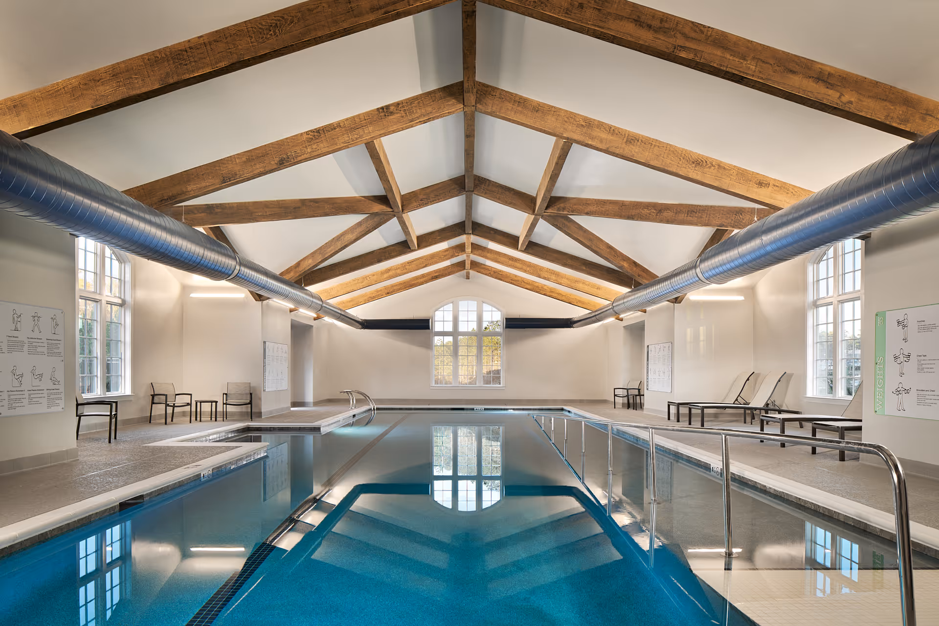 Indoor swimming pool with clear blue water in a room featuring high vaulted ceilings with exposed wooden beams and large windows. The pool area has metal handrails, lounge chairs, and chairs along the sides. Exercise instruction posters are mounted on the walls.