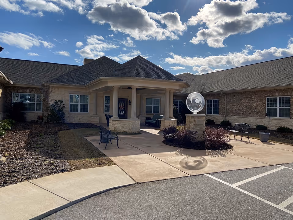 Front entrance of a single-story brick senior living building with a covered portico, outdoor benches, and a glass orb sculpture at the driveway.