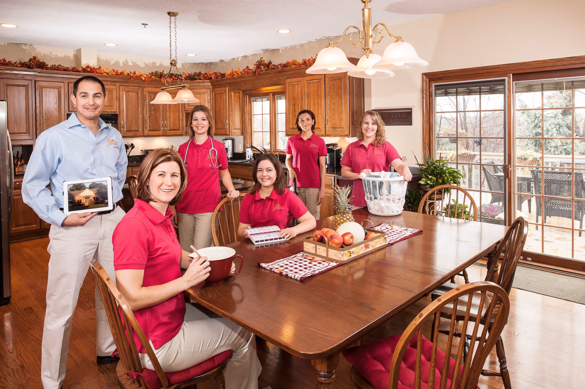 A group of six smiling staff members in a kitchen and dining area. Five women wearing matching red polo shirts and beige pants, one man in a light blue shirt holding a tablet displaying a building photo. The kitchen has wooden cabinets, a large wooden dining table with fruit centerpiece, and sliding glass doors leading to an outdoor patio.