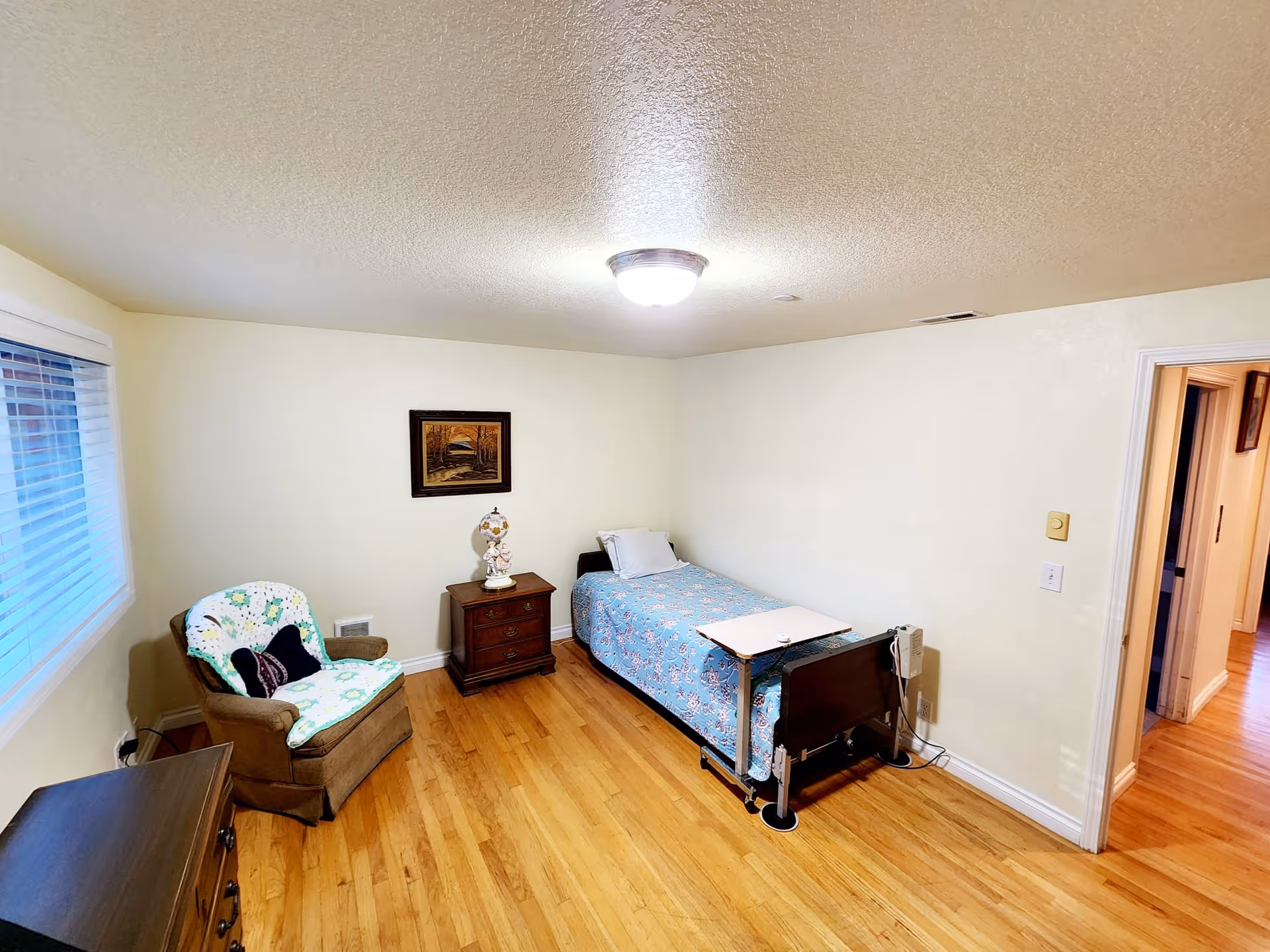 A small bedroom with a single bed covered in a blue floral bedspread, a wooden nightstand with a decorative lamp, a cushioned armchair with a floral cover and a black pillow, a wooden dresser, a window with blinds, and a framed painting on the wall. The floor is hardwood, and the room is lit by a ceiling light.