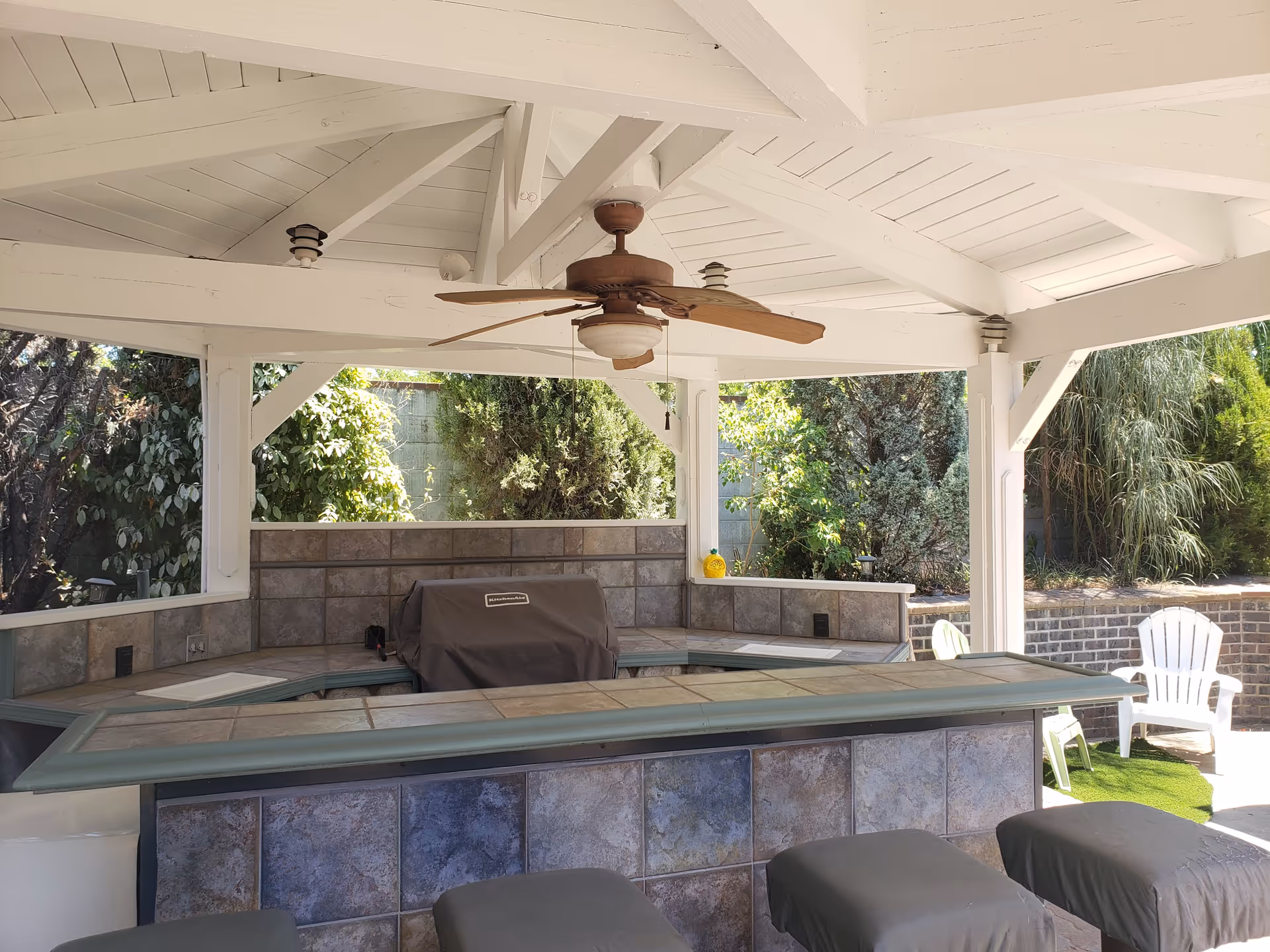 Outdoor covered patio area with a ceiling fan, tiled bar counter with stools, a covered grill, and greenery in the background.