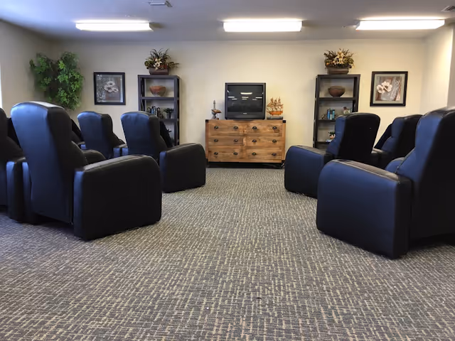 A senior living facility common area with eight black recliner chairs arranged in two rows facing a wooden dresser with a television on top. Two black shelving units flank the dresser, each decorated with plants, pottery, and books. The room has carpeted flooring, neutral-colored walls, and ceiling lights.