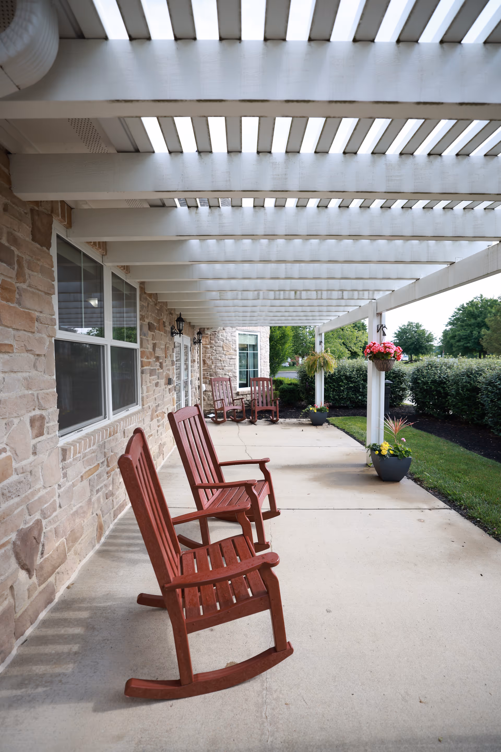 Covered patio with red wooden rocking chairs along a stone building under a white pergola with hanging flower pots.