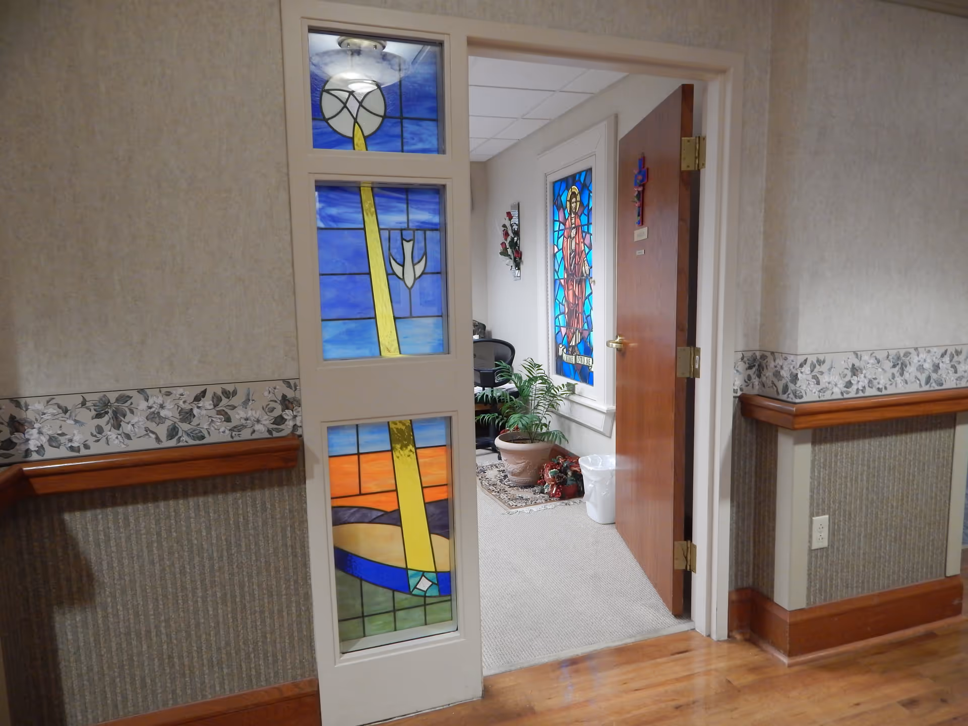 Interior hallway view showing an open wooden door with a stained glass panel depicting a religious figure. Next to the door is a wall with three vertical stained glass panels featuring abstract designs in blue, yellow, and orange colors. Inside the room, there is a potted plant on a small rug, a chair, and a white trash bin. The hallway walls have floral wallpaper border and wooden handrails.