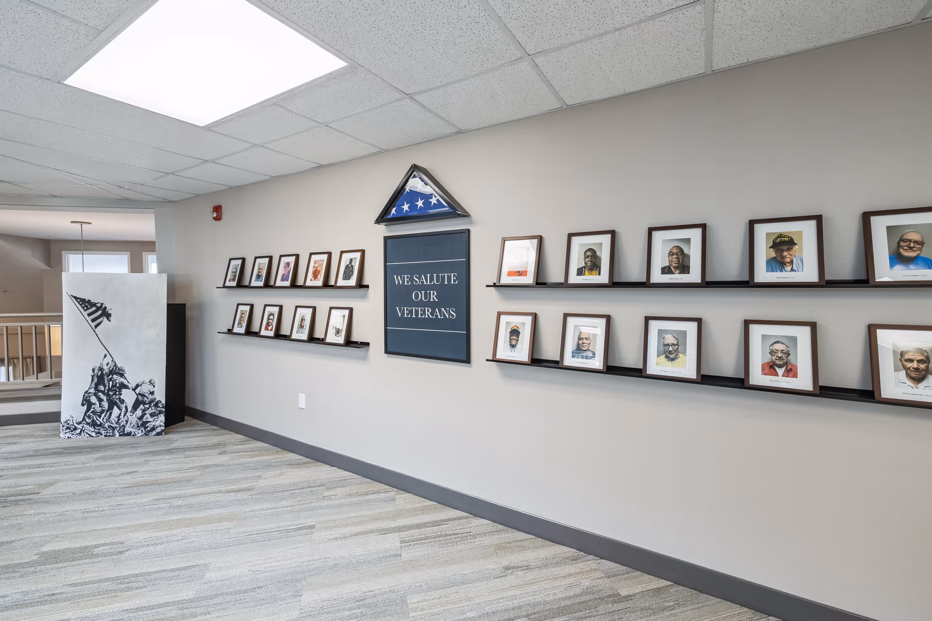 Interior hallway with a display honoring veterans. The wall features framed photographs of veterans arranged on two black shelves, a folded American flag in a triangular display case, and a sign that reads 'WE SALUTE OUR VETERANS'. To the left, there is a black and white image of soldiers raising an American flag. The floor is carpeted in a light gray pattern and the ceiling has white tiles with fluorescent lighting.