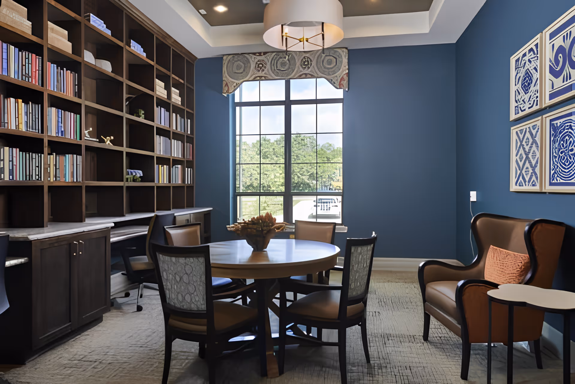 A cozy interior room with a round wooden table surrounded by four chairs, a large window with a decorative valance letting in natural light, a tall bookshelf filled with books and decorative items, a comfortable brown armchair with an orange pillow, and blue walls adorned with four framed blue and white artworks.