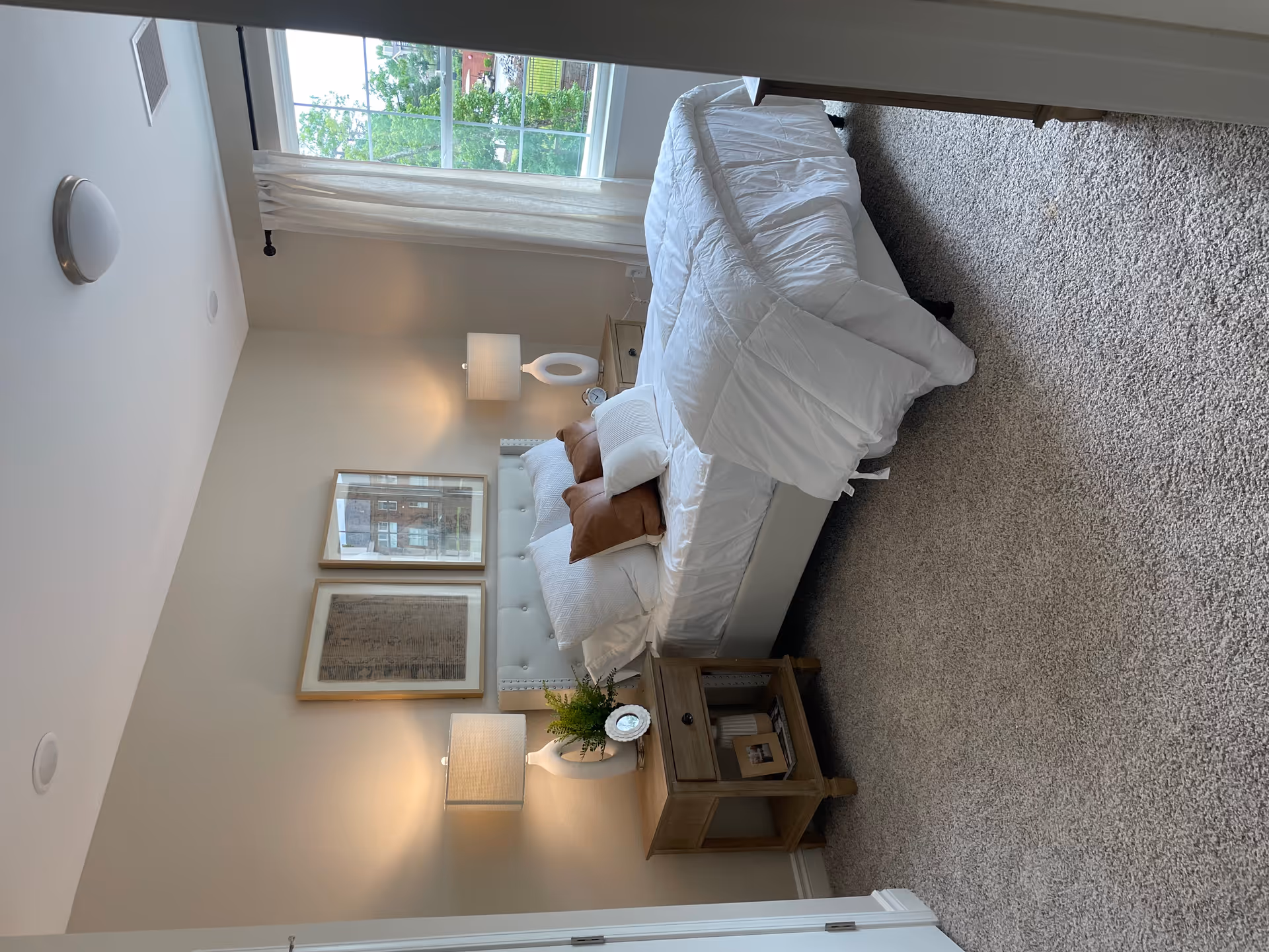 Furnished bedroom with a white-made bed, tufted headboard, two wooden nightstands with lamps, framed artwork and a window.